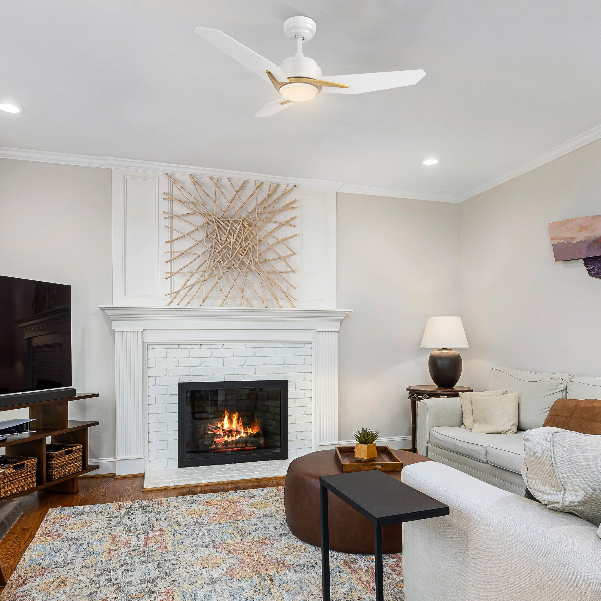 A cozy living room with a Bryza 44-inch white and gold ceiling fan with light, a burning white brick fireplace, a flat-screen TV, a light gray sofa with cushions, a side table with a lamp and plant, a brown ottoman with a tray, and a black side table over a colorful rug on a wooden floor. 