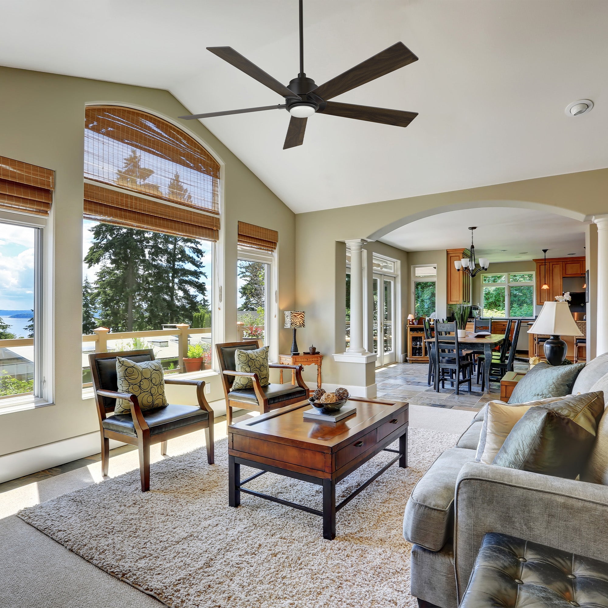 Kalmar 60-inch remote control ceiling fan with LED light installed in a bright living room featuring large windows, wooden furniture, and an open-concept layout connecting to the dining area. 