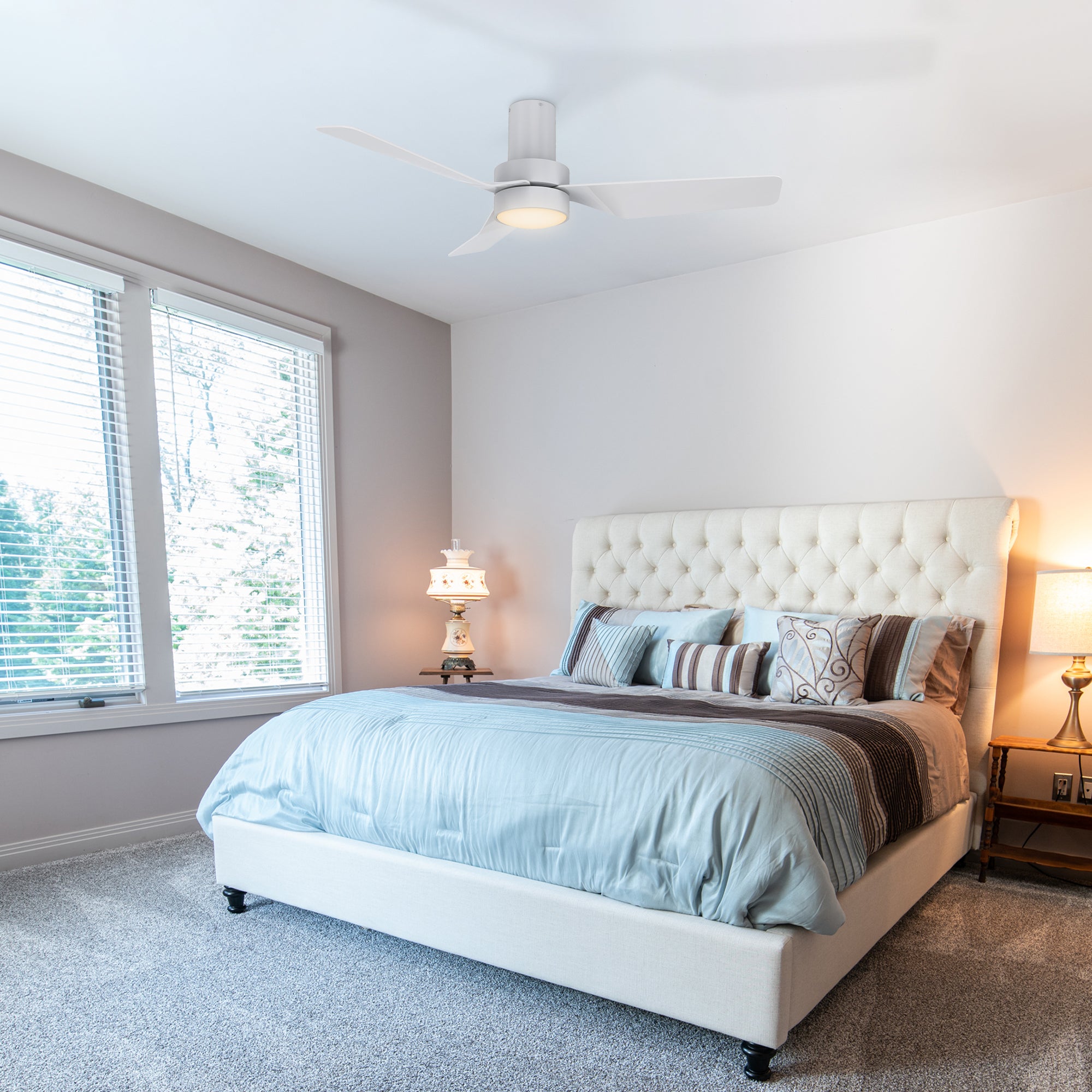 A bedroom featuring a Smafan Carro Marion 44-inch remote-controlled white ceiling fan with a light kit. The room has a tufted headboard, blue and beige bedding, vintage nightstand lamps, light grey walls, carpeted floors, and large windows with white blinds. #color_white