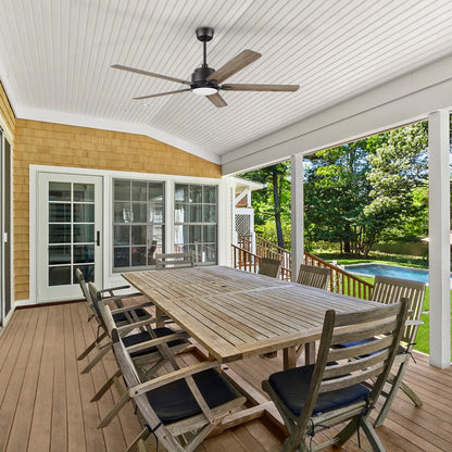 A spacious covered patio with a long wooden dining table and eight chairs, featuring the 56-inch Smafan Essex outdoor smart ceiling fan in walnut finish. The fan is mounted on a white paneled ceiling, providing cooling and lighting for the outdoor dining area. The space overlooks a backyard with lush greenery and a swimming pool. 