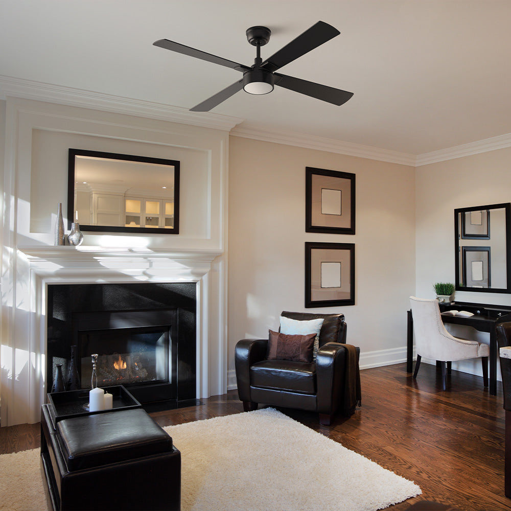 A Smafan Admiral 52-inch matte black 4-blade ceiling fan with a circular LED light, installed in a modern living room. The room features a white fireplace surround with a black marble hearth and a burning fire, a dark brown leather armchair, dark hardwood flooring, and a white area rug. 