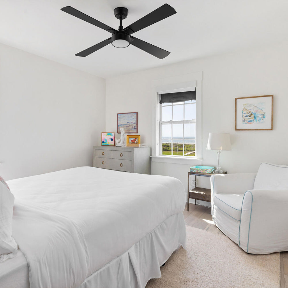 A Smafan Admiral 52-inch matte black ceiling fan with an integrated frosted LED light kit, installed on a white bedroom ceiling. The bright room features a bed with white bedding, a light gray dresser with colorful framed art, a white armchair, and a window showing a coastal view. 