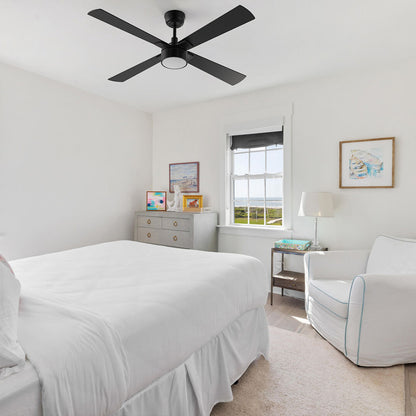 A Smafan Admiral 52-inch matte black ceiling fan with an integrated frosted LED light kit, installed on a white bedroom ceiling. The bright room features a bed with white bedding, a light gray dresser with colorful framed art, a white armchair, and a window showing a coastal view. 