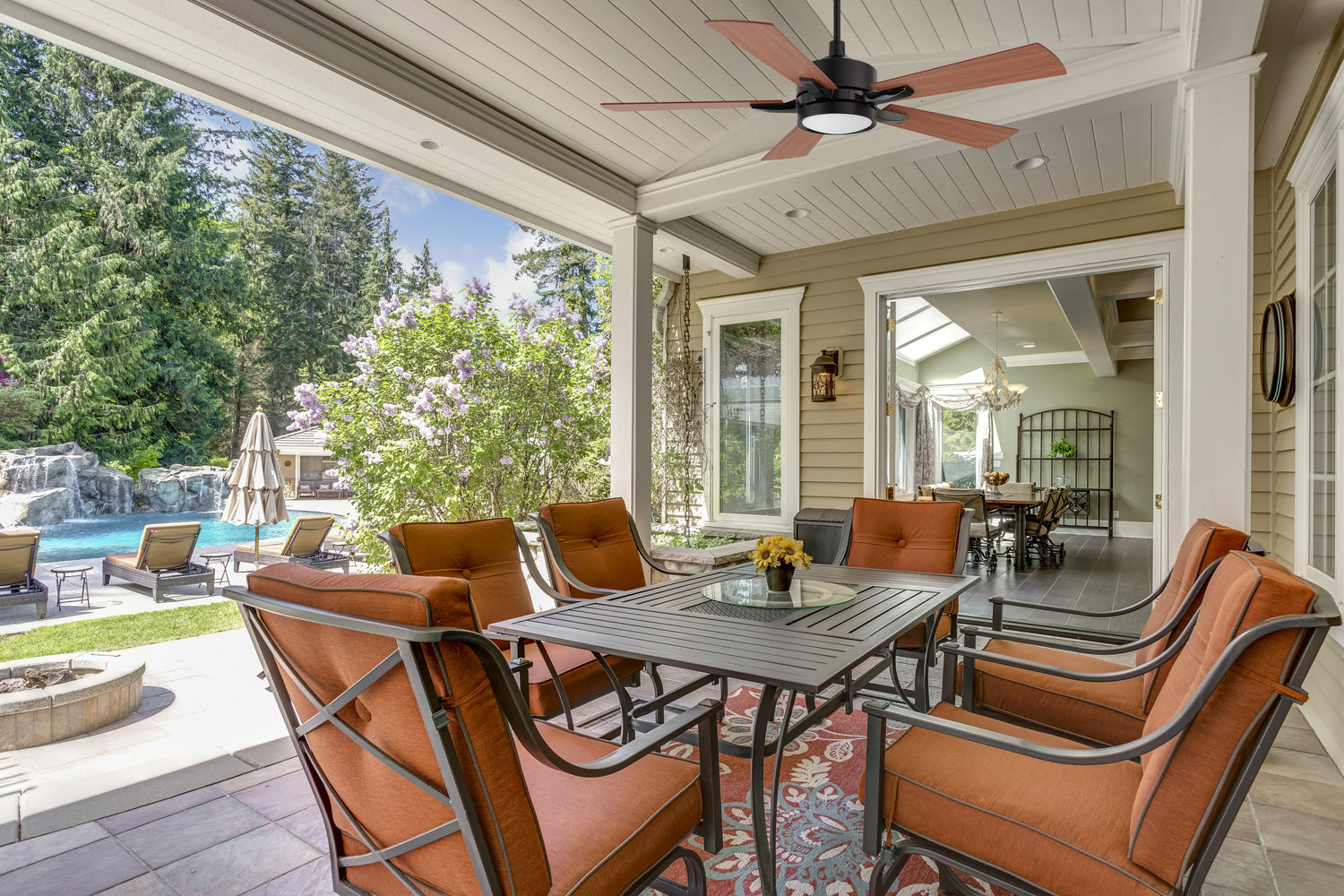 A covered patio with cushioned orange chairs around a square table features an outdoor smart ceiling fan with light above, and a view into a living area. In the background, there’s a pool, lounge chairs, and green trees.