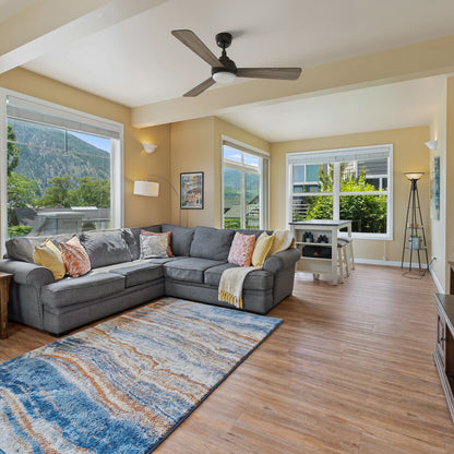 A bright modern Bohemian living room with a gray sectional sofa, colorful pillows, and a yellow throw, centered around a blue and orange area rug on warm wood flooring. Overhead, the Smafan Audra 52-inch 3-blade ceiling fan with light provides airflow and illumination. Large windows frame mountain views, enhancing the inviting space. 