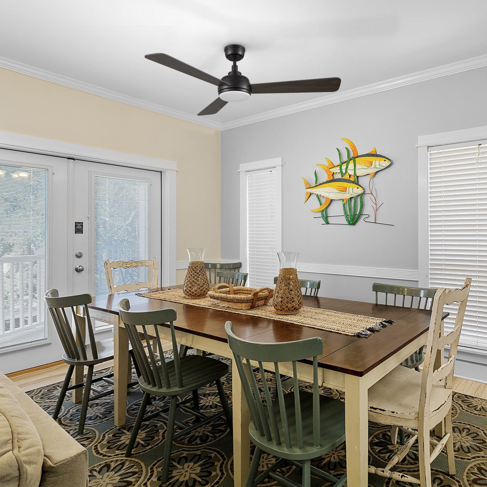 A rustic dining room features a long wooden table with mismatched chairs in shades of green and cream. Above the table hangs a Smafan Audra 52-inch 3-blade ceiling fan with a light, in a dark wood finish. The room has light-colored walls, large windows with blinds, and a patterned rug. Decorative fish wall art adorns one wall. 