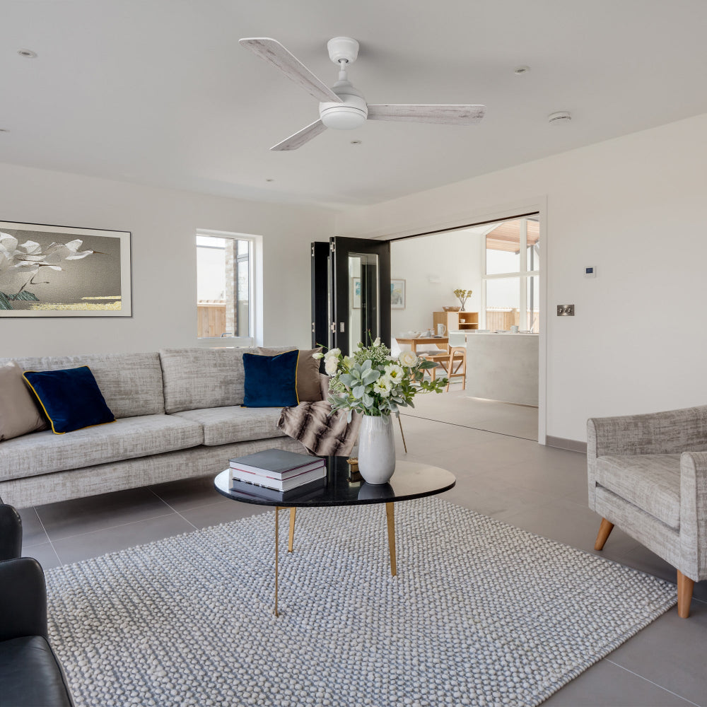 A bright modern living room with a Smafan Audra 52-inch white ceiling fan with light mounted on the ceiling. The 3-blade fan features a minimalist design and integrated LED. The space includes a gray sofa with blue and beige pillows, a round black coffee table with flowers and books, a gray rug, and large windows bringing natural light. 