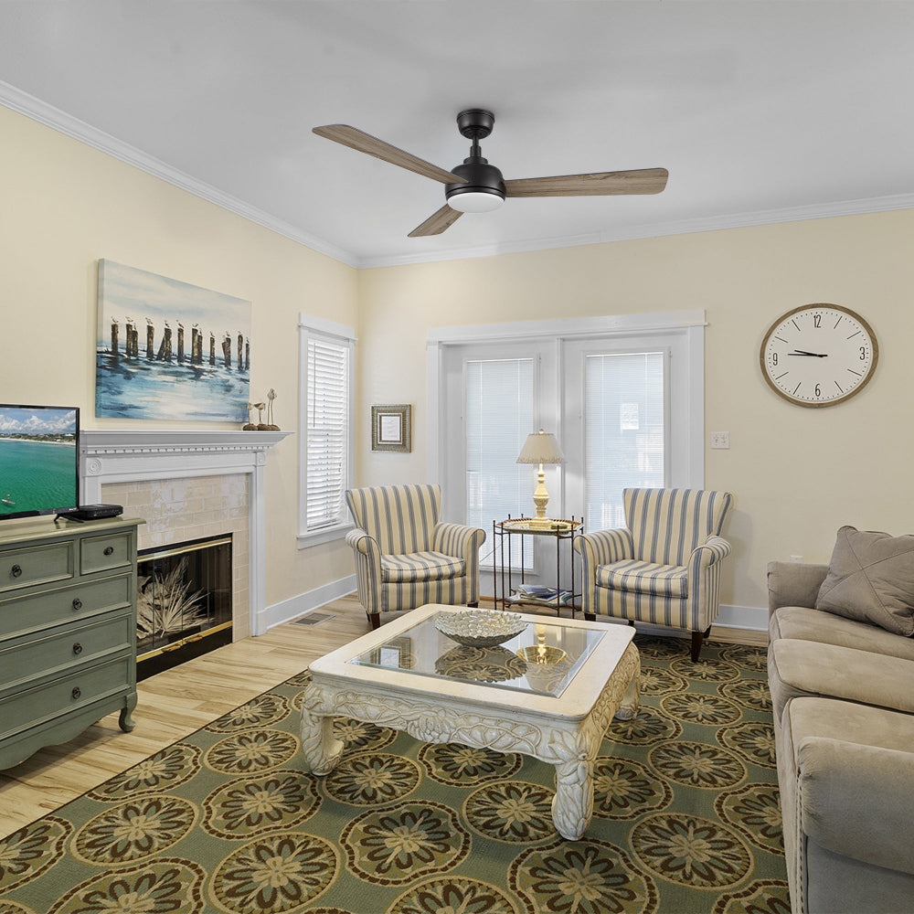 A Modern Traditional living room featuring a Smafan 52 inch Audra wood ceiling fan with light. The room includes a beige sofa, two striped armchairs, a detailed white coffee table with a glass top, and a fireplace with a TV and a large abstract painting above it. The space is anchored by a patterned area rug and illuminated by natural light from large windows with blinds, as well as a table lamp on a side table between the armchairs 