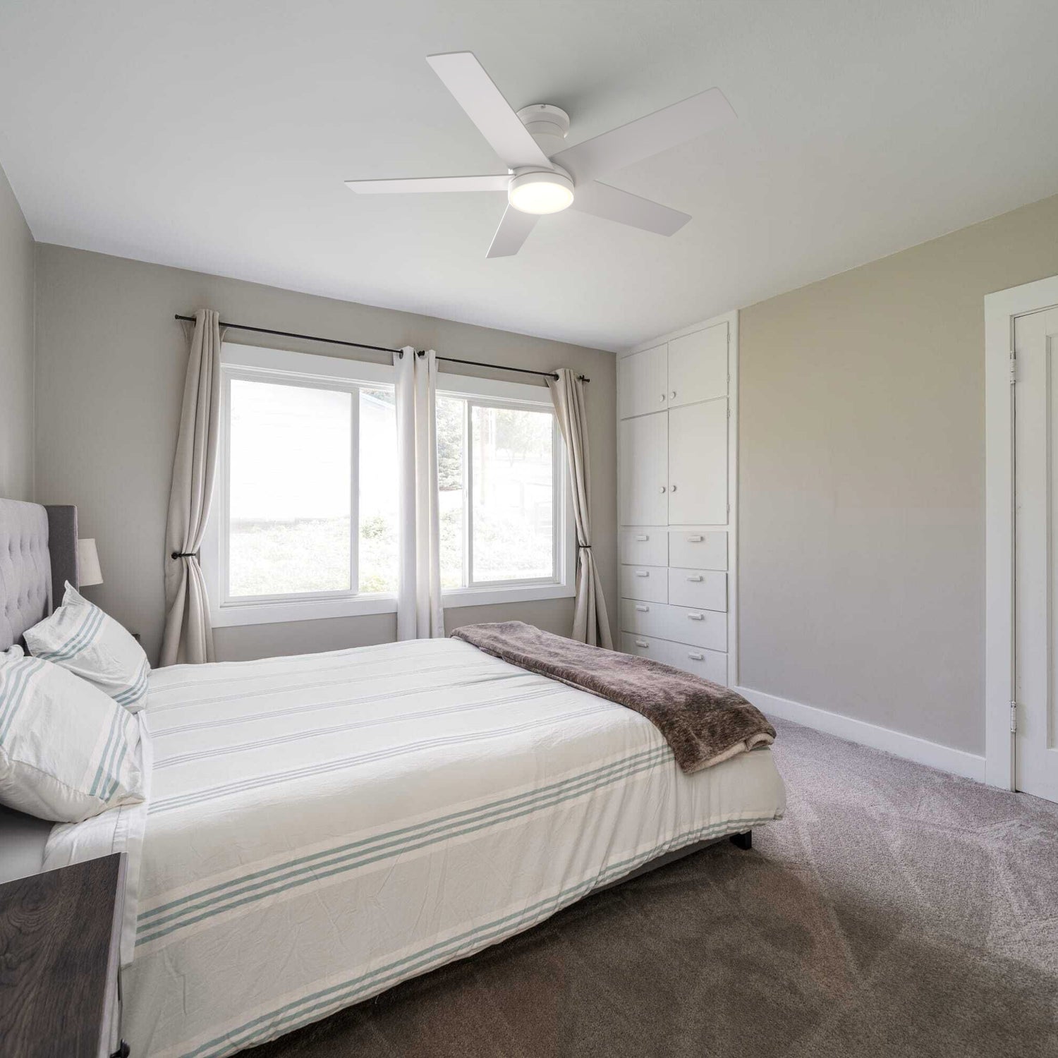 Modern bedroom with white Bayu 52 inch low profile ceiling fan with LED light, white comforter with green stripes, built-in wardrobe, and large windows with sheer curtains. 
