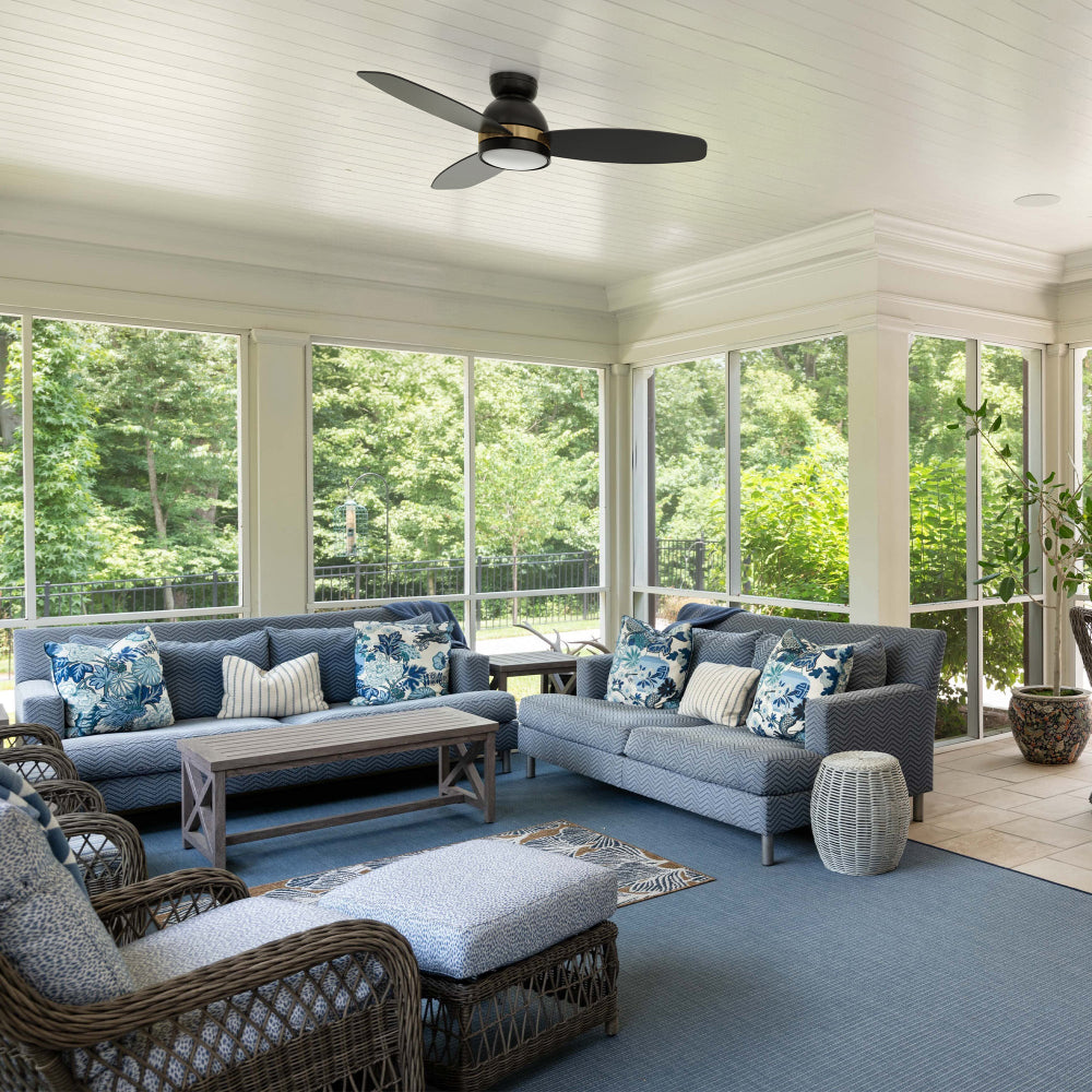 A screened-in porch with blue and white patterned furniture, including two sofas, a coffee table, and wicker chairs, under a white coffered ceiling with a black and gold Smafan Biscay 48-inch low-profile ceiling fan with light, overlooking a lush green outdoor area 