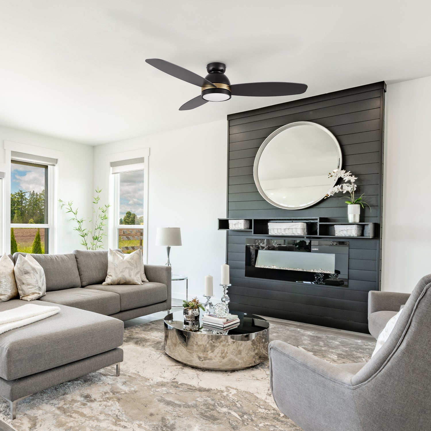 Modern living room featuring the Smafan Biscay 48-inch smart ceiling fan in black with integrated light. The space includes a grey sectional sofa, round mirrored coffee table, and a black accent wall with a circular mirror and electric fireplace. 