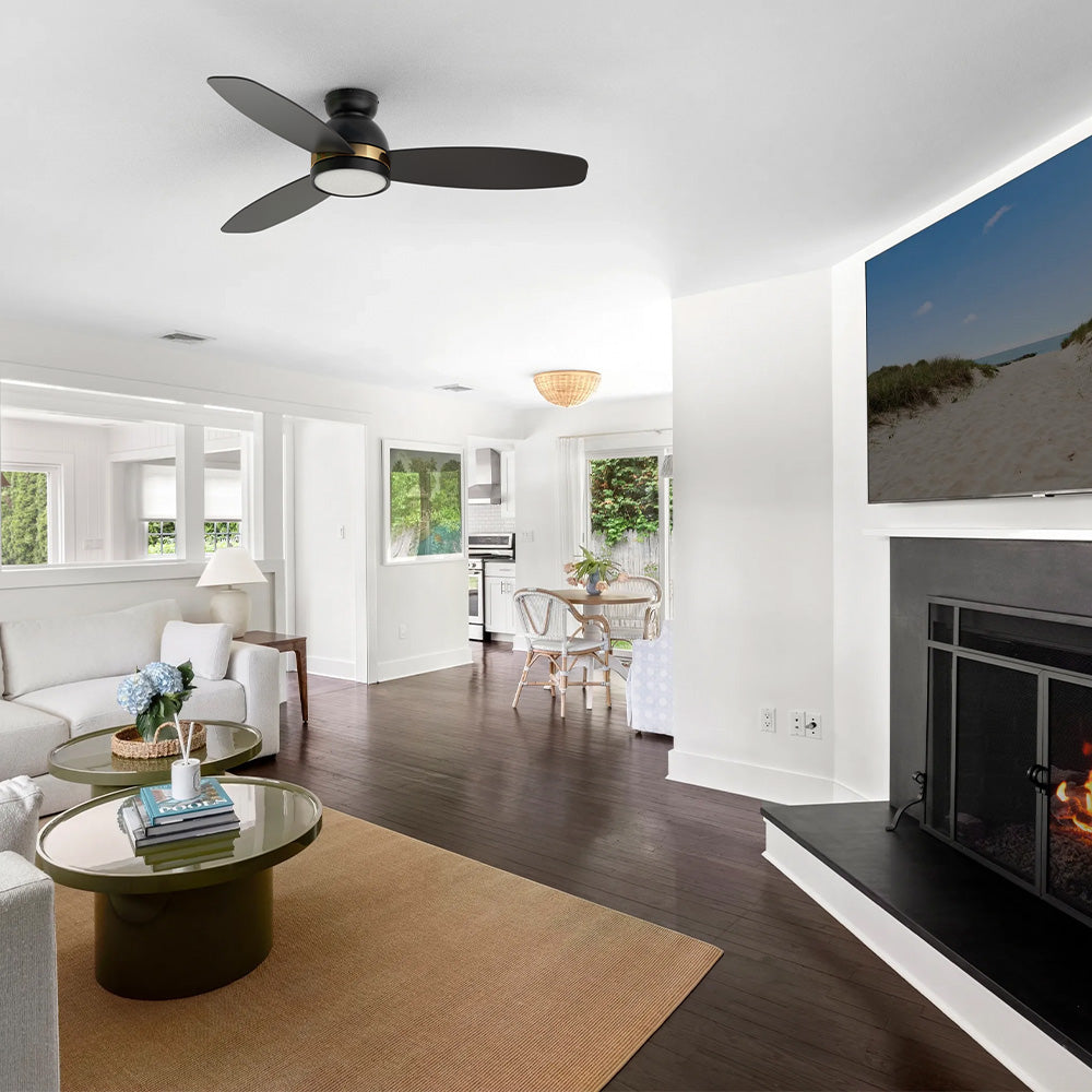 Black and gold flush mount 3-blade ceiling fan with LED light installed on the ceiling of a bright open concept living room and dining area with a fireplace. 