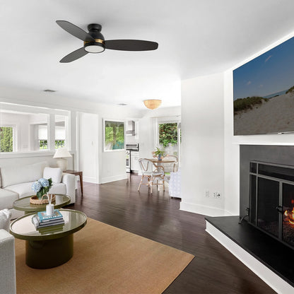 Black and gold flush mount 3-blade ceiling fan with LED light installed on the ceiling of a bright open concept living room and dining area with a fireplace. 