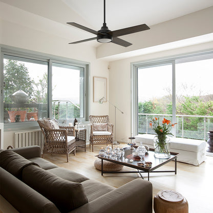 Smafan Bryant black ceiling fan with light installed in a sunlit coastal cottage transitional living room with wicker armchairs, large glass windows, and a brown sofa. 