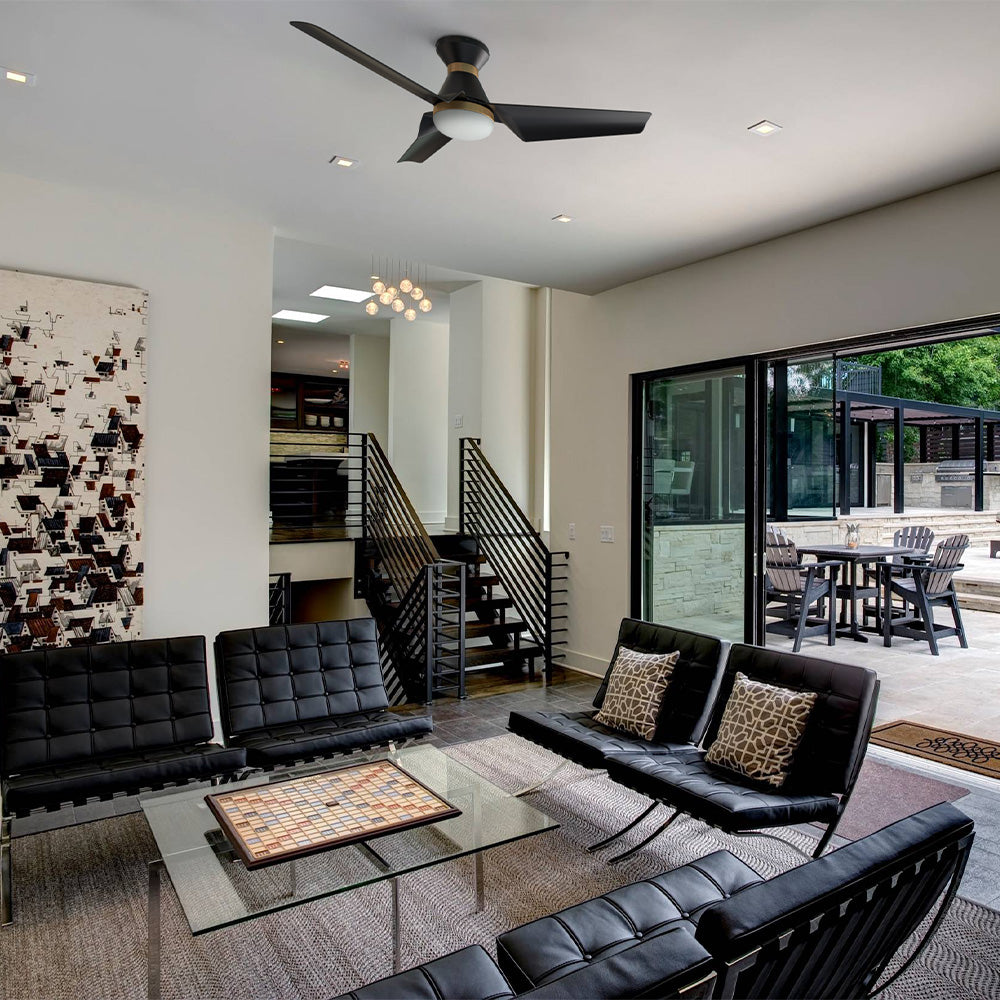A modern living room featuring black leather seating, a glass coffee table with a board game, an abstract painting, a flush mount ceiling fan in black and gold, and large sliding glass doors leading to an outdoor patio area. 