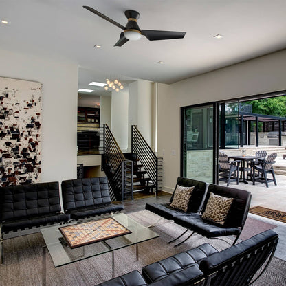 A modern living room featuring black leather seating, a glass coffee table with a board game, an abstract painting, a flush mount ceiling fan in black and gold, and large sliding glass doors leading to an outdoor patio area. 
