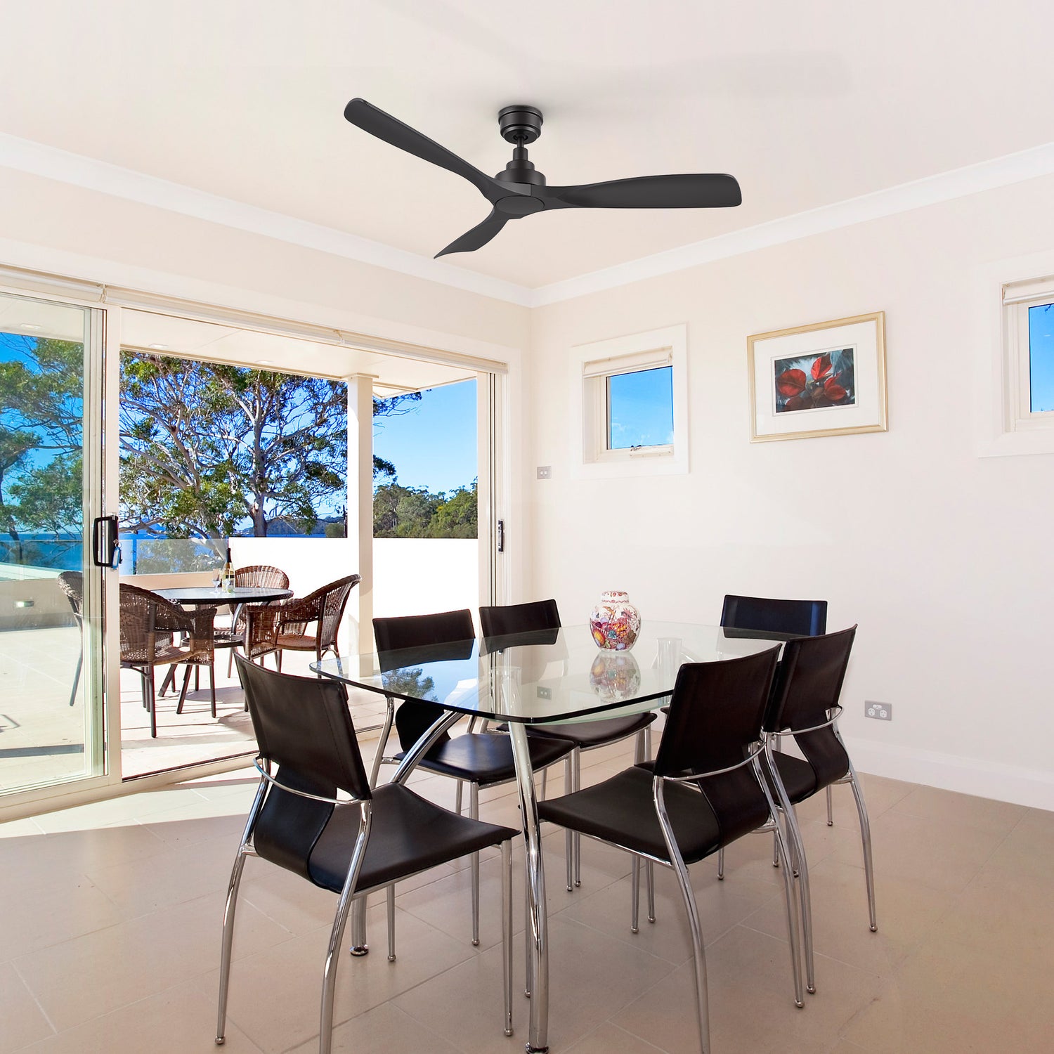 A living room featuring a 52-inch Smafan Kilk smart ceiling fan in black, without light, mounted on a white ceiling. The room includes a glass dining table with six black leather chairs, a colorful vase as a centerpiece, and light beige walls with a framed picture. Large sliding glass doors open to a balcony with wicker furniture and a view of trees under a clear blue sky, with tiled flooring throughout. 