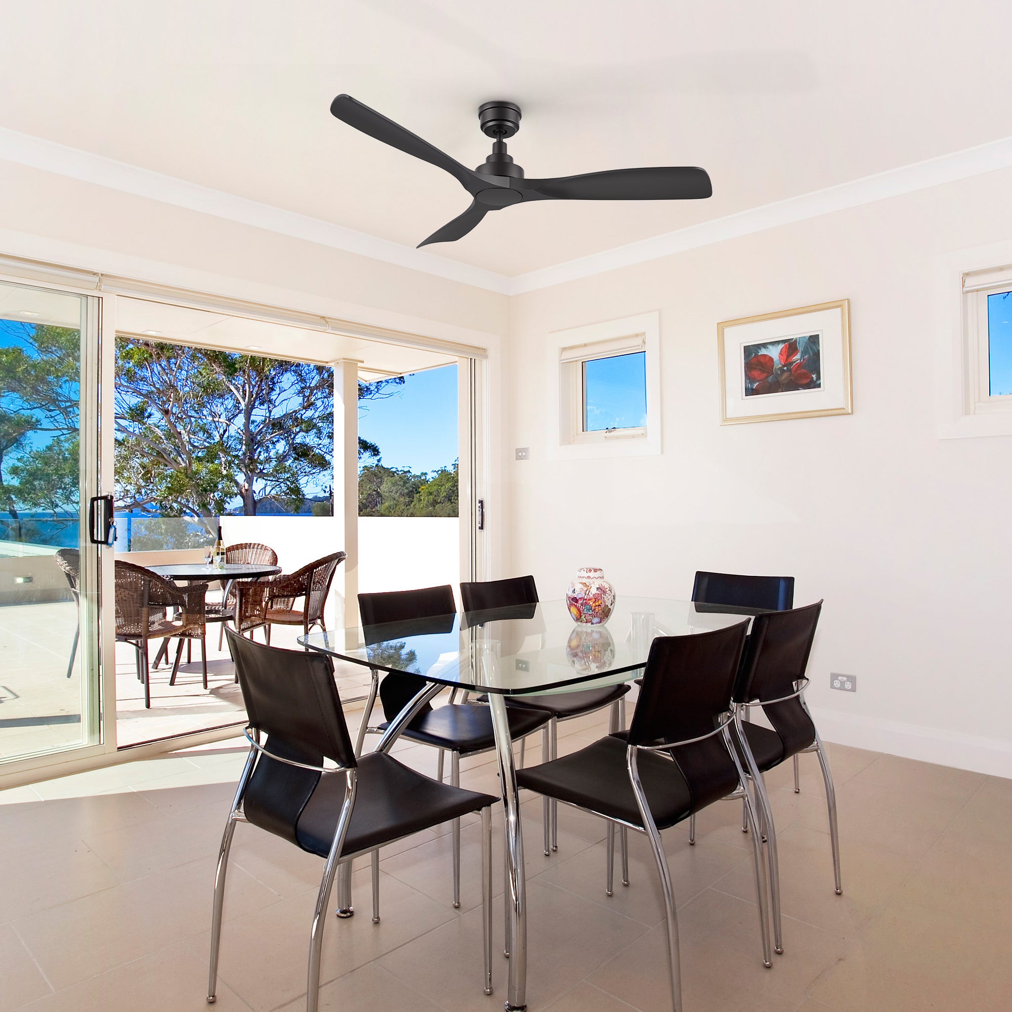 A living room featuring a 52-inch Smafan Kilk smart ceiling fan in black, without light, mounted on a white ceiling. The room includes a glass dining table with six black leather chairs, a colorful vase as a centerpiece, and light beige walls with a framed picture. Large sliding glass doors open to a balcony with wicker furniture and a view of trees under a clear blue sky, with tiled flooring throughout. 