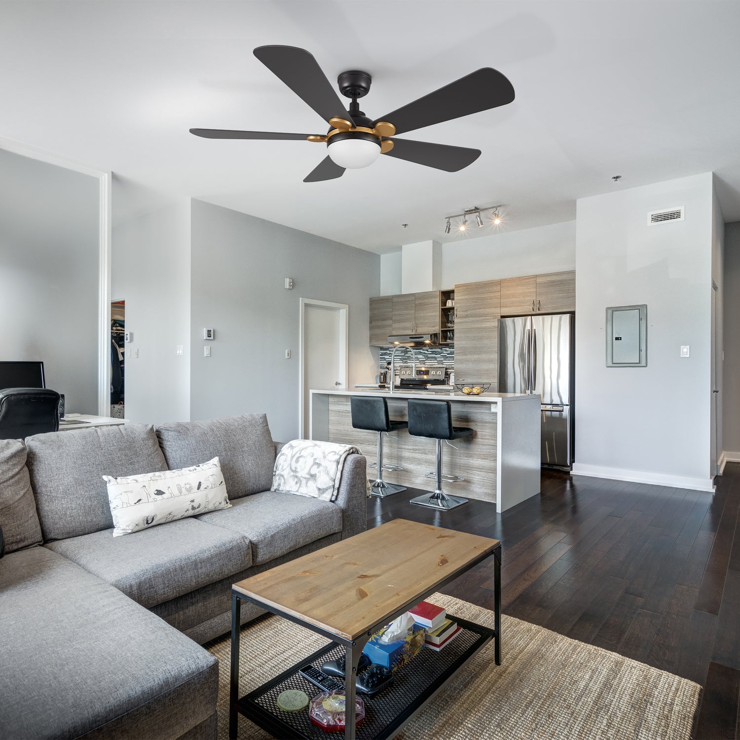 Smafan Lille 52 inch unique ceiling fan in black and gold with light fixture is centered in a modern living room and kitchen area. The living room features a large gray sectional sofa with a patterned pillow and a throw blanket, a wooden coffee table with a metal base, and a woven rug. The kitchen, visible in the background, has a white island with two black bar stools, light wood cabinetry, a stainless steel refrigerator, and tiled backsplash. 