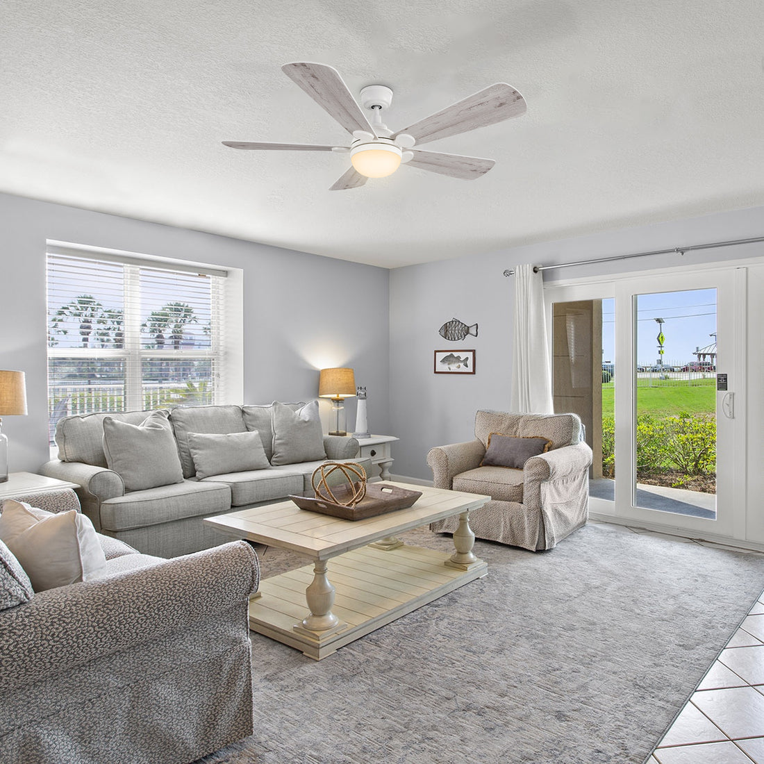 A bright and airy living room with a light gray sofa and two armchairs, a white coffee table, a light-colored area rug, and 52 inch Smafan Lille white ceiling fan with bright light and light wood colored blades. A large window with blinds is on the left, and a glass sliding door leading to an outdoor grassy area is on the right. Decorative fish-themed artwork adorns the wall. 