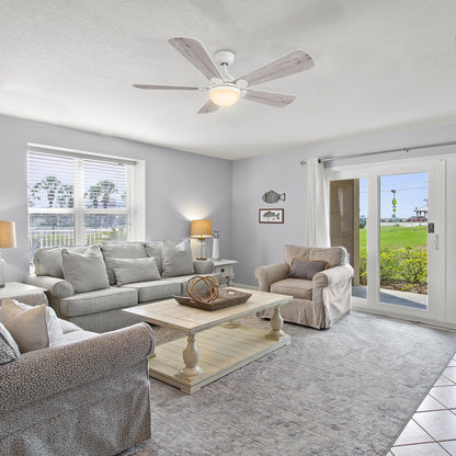 A bright and airy living room with a light gray sofa and two armchairs, a white coffee table, a light-colored area rug, and 52 inch Smafan Lille white ceiling fan with bright light and light wood colored blades. A large window with blinds is on the left, and a glass sliding door leading to an outdoor grassy area is on the right. Decorative fish-themed artwork adorns the wall. 