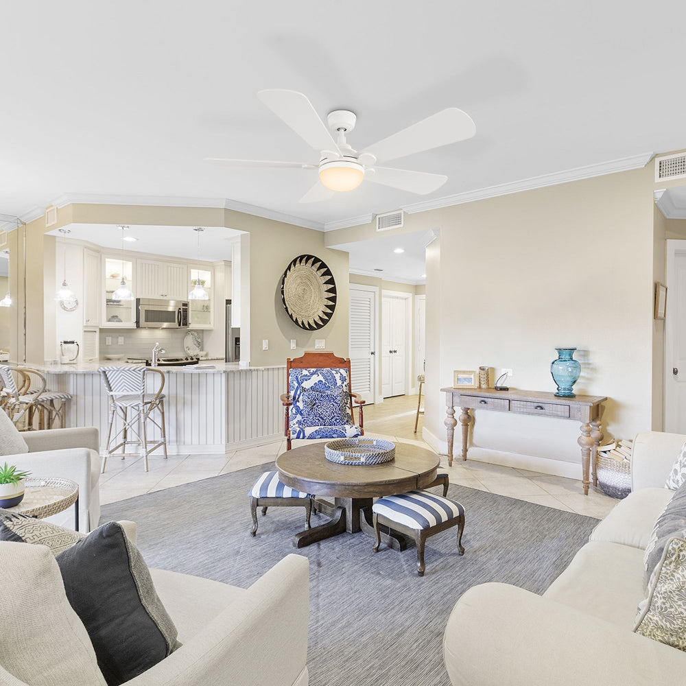 A bright and airy living room and kitchen area in a coastal-themed home, featuring white furniture, 52 inch Smafan Lille large ceiling fan with bright light in warm tone, a round wooden coffee table with striped ottomans, and a kitchen island with bar stools. 
