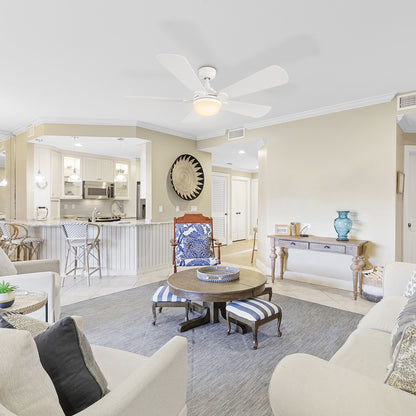 A bright and airy living room and kitchen area in a coastal-themed home, featuring white furniture, 52 inch Smafan Lille large ceiling fan with bright light in warm tone, a round wooden coffee table with striped ottomans, and a kitchen island with bar stools. 