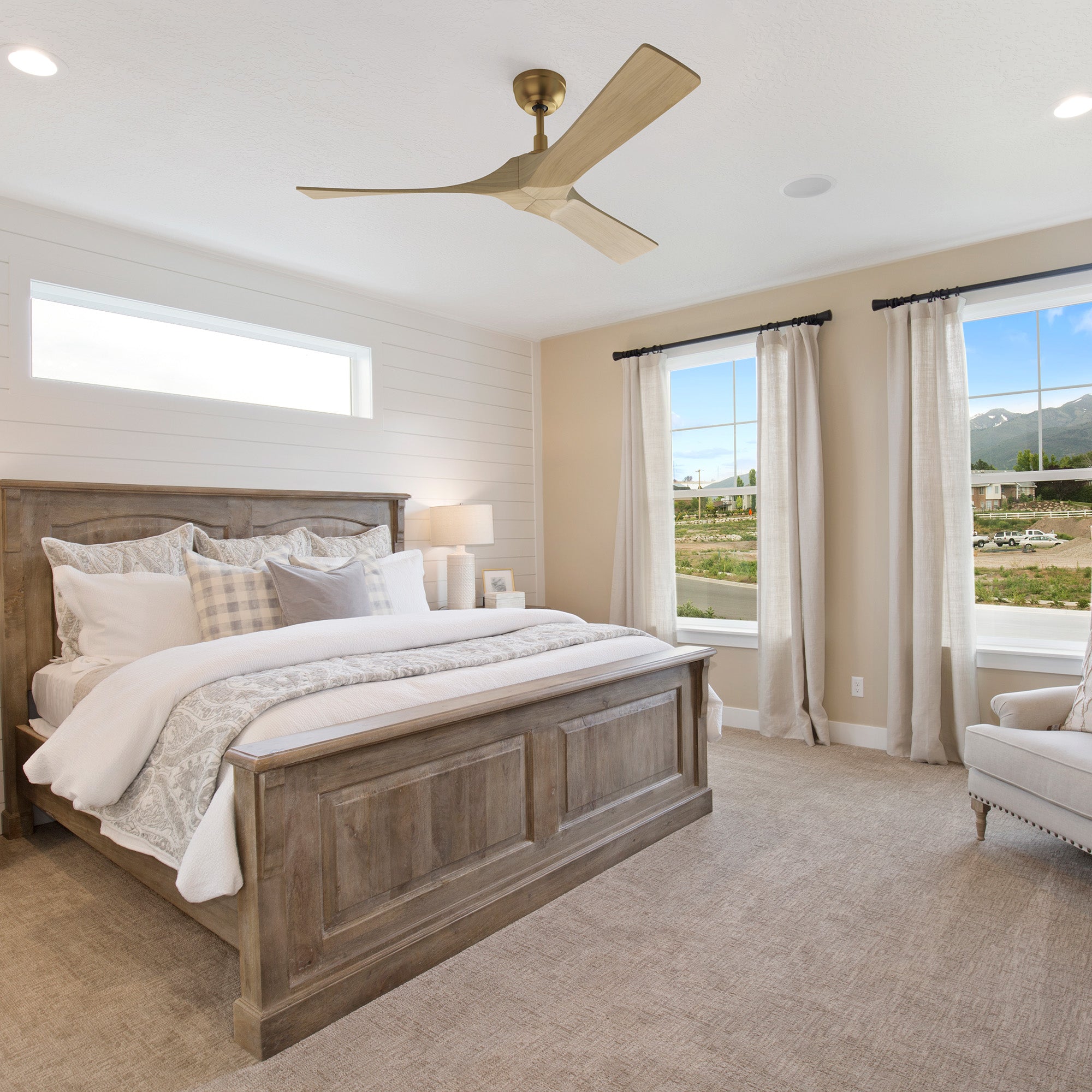 A bedroom featuring a 52-inch Smafan Matthews brass ceiling fan with remote and no LED light, mounted on the ceiling. The room includes a wooden bed with a white and gray patterned comforter, multiple pillows, and nightstands with lamps. The space has light beige walls with shiplap detailing, large windows with white curtains offering a view of mountains and greenery, carpeted flooring, and a beige armchair in the corner. 