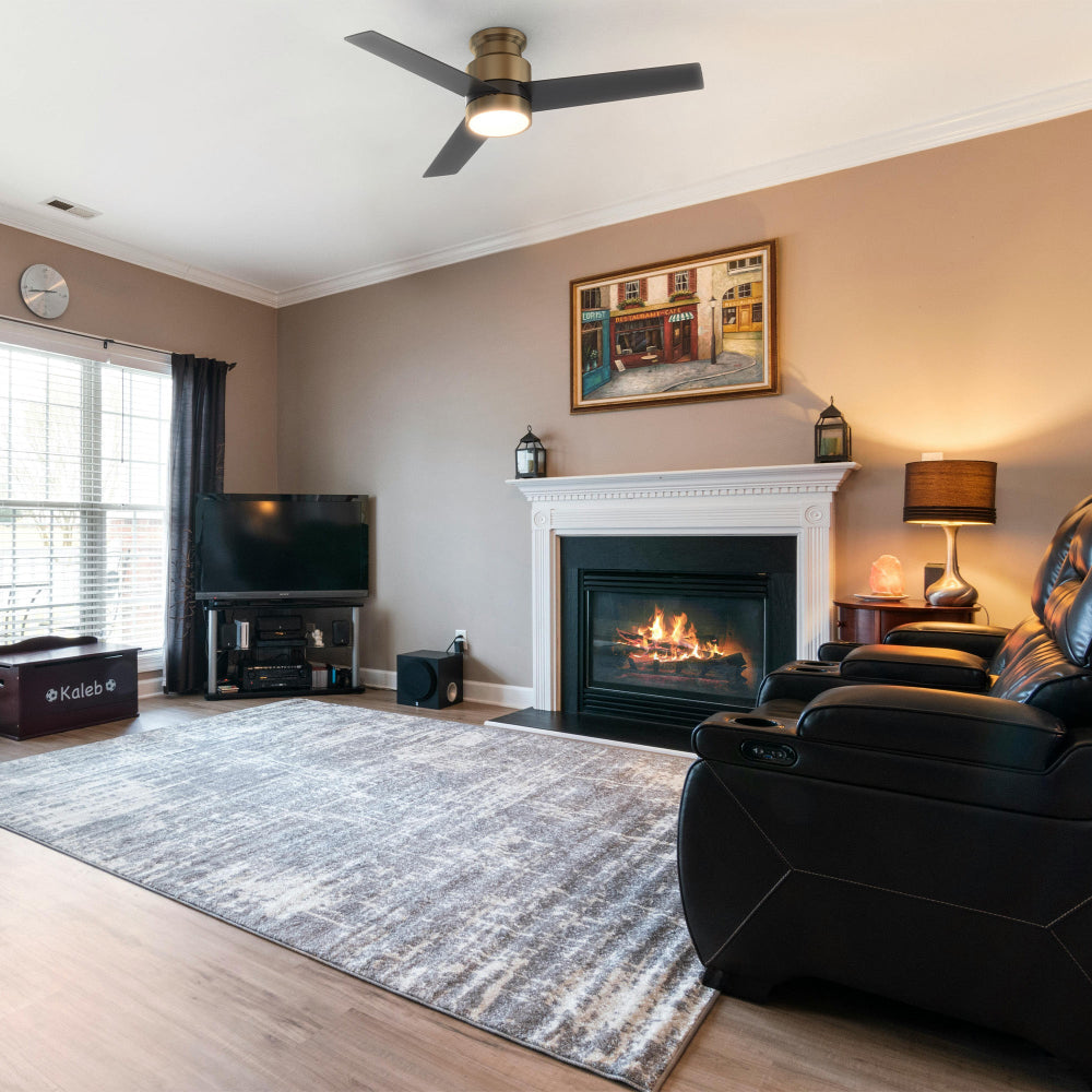 A modern living room with fireplace and leather recliners featuring the Smafan Ranger Smart Ceiling Fan in brushed gold / brass with black blades. This low profile ceiling fan is ideal for large indoor living spaces, equipped with an energy-efficient LED light kit that provides soft illumination to the neutral-toned decor. 