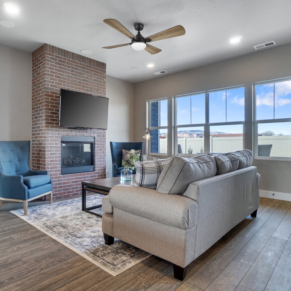 A living room featuring a fireplace with a mounted television above it, a modern black ceiling fan with light kit, a light-colored sofa, two blue armchairs, a coffee table, and large windows providing a view of the outdoors. 