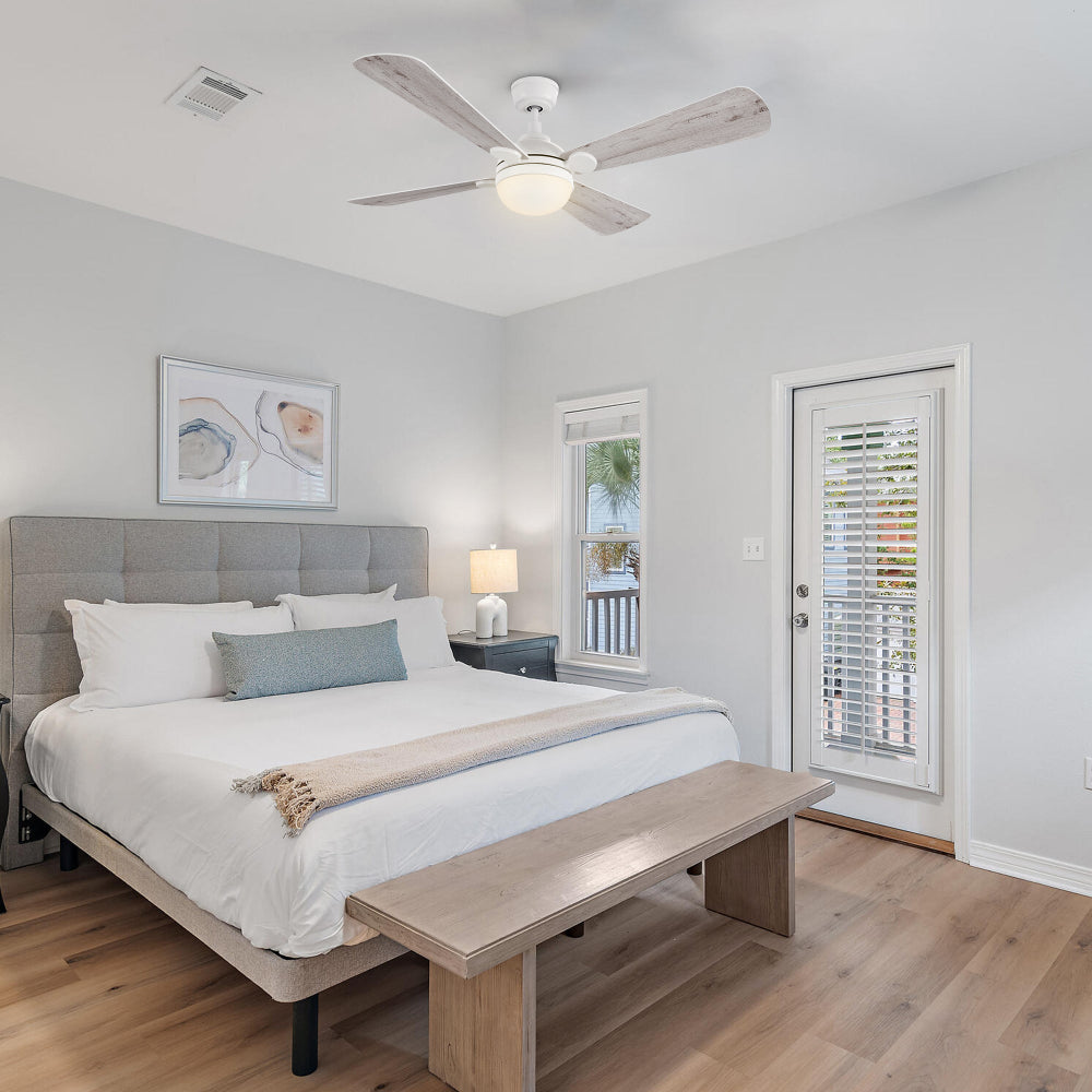 A bedroom featuring a Smafan Rinard 52-inch white ceiling fan with light, a large bed with a gray upholstered headboard, a wooden bench at the foot of the bed, and a door leading to an outdoor space. The room has an eclectic style with light-colored walls and wood flooring. 