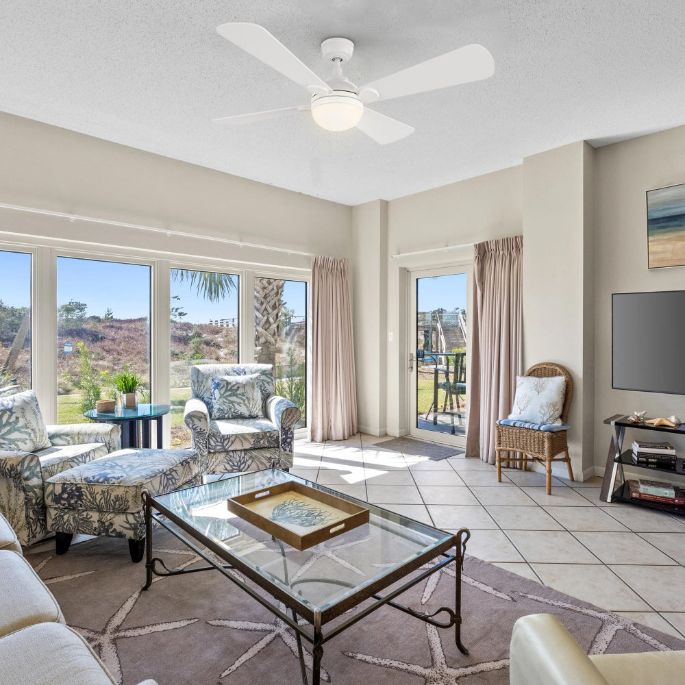 A white Smafan Rinard ceiling fan with light is installed in a traditional-style living room, which features large windows overlooking a green landscape, patterned armchairs, a glass coffee table with a tray, and a starfish-patterned rug.  