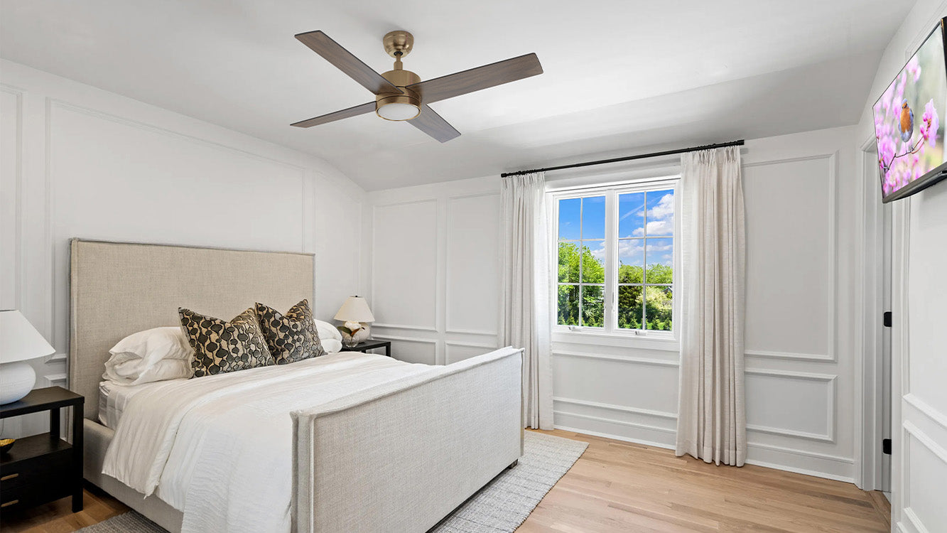 Bedroom scene with a gold ceiling fan with light and wooden blades mounted on a white ceiling at the top. The room features white walls with decorative paneling and a large window showing green trees and a blue sky. The bed has a white headboard with black-patterned pillows and white bedding, next to a black nightstand with a white lamp. A TV on the right wall displays pink flowers, and the floor is wooden.