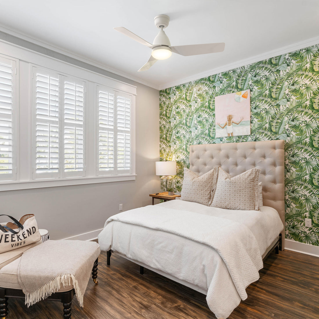 A modern bedroom featuring a Smafan Silas 44-inch Modern Smart Ceiling Fan in white with Light and Remote on the ceiling, a bed with a tufted headboard and white bedding, a nightstand with a lamp, and a bench with a blanket and a &quot;WEEKEND VIBES&quot; tote bag. The room has light gray walls and a vibrant green and white palm leaf patterned accent wall behind the bed, with white plantation shutters on the windows. 