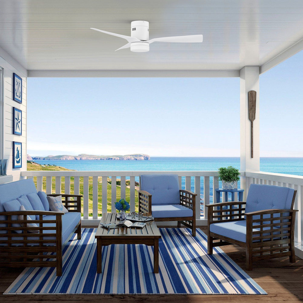 Smafan Striver II low-profile white smart ceiling fan with light installed on a covered balcony overlooking the ocean. The coastal patio features blue cushioned wooden chairs, a striped rug, and decorative nautical accents. Ideal for outdoor covered spaces. 