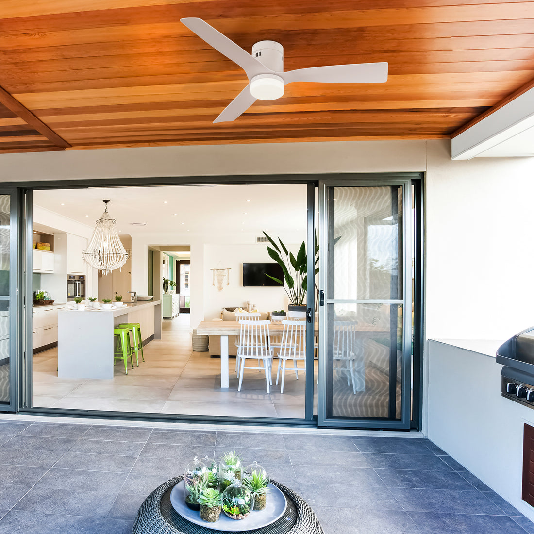 A covered balcony featuring a 52-inch Smafan Striver2 outdoor smart ceiling fan in white with a light, mounted on a wooden ceiling. The balcony has a gray tiled floor with a small table displaying terrariums with succulents. Sliding glass doors open to an indoor space with a modern kitchen featuring white cabinets, green stools, a chandelier, and a dining area with a wooden table and white chairs, visible in the background. 