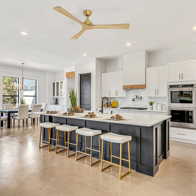 Smafan Thora 60 inch wood ceiling fan with remote in a gold finish, mounted above a large gray kitchen island with white countertop and gold bar stools. The modern open-concept kitchen features white cabinets, stainless steel appliances, a wood range hood, and a dining table in the background. #color_gold