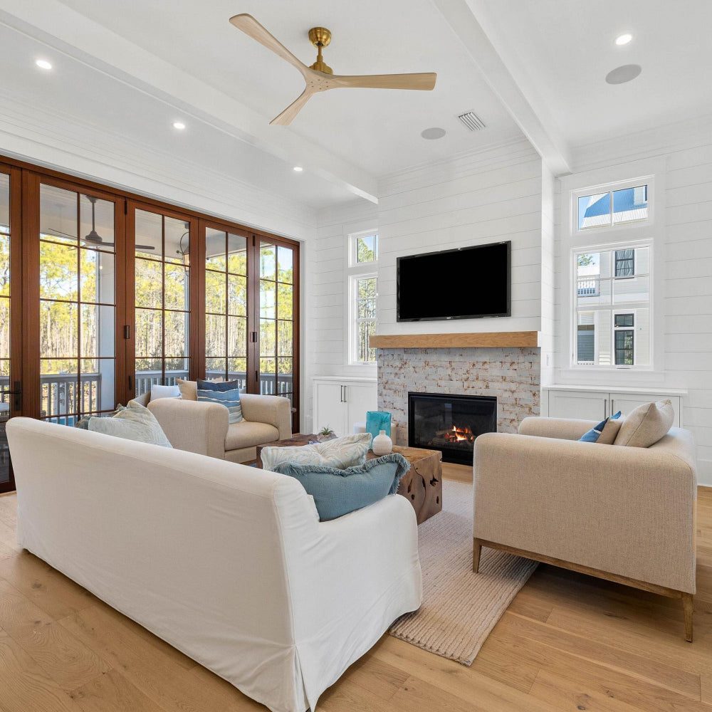 Smafan Thora 60 inch wood ceiling fan with remote in a gold finish, installed in a bright modern farmhouse living room with white shiplap walls, a fireplace with flat-screen TV above, light-colored sofas and armchairs around a wooden coffee table, and large glass doors opening to an outdoor area. 