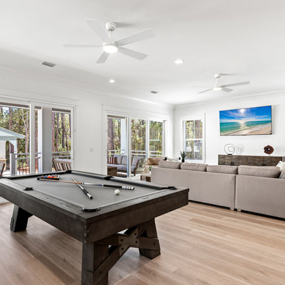 A modern open-concept living space featuring two Smafan Toplam 52-inch white ceiling fans with lights on the ceiling. The room includes a dark wood pool table with a gray felt top, a large beige sectional sofa facing a wall-mounted TV, and expansive glass doors revealing a forest-view deck with patio furniture. 