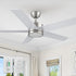 A modern 52-inch Smafan Trafford2 brushed nickel ceiling fan with light, featuring five white blades, installed in a bright room with beige curtains and a roman shade. Two control ways are shown below: a wall control labeled "CARRO" with multiple buttons for fan and light settings, and a handheld remote with similar functionality. 