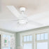 A modern 52-inch Smafan Trafford2 white ceiling fan with a 
 light, featuring five white blades, installed in a bright room with large windows and white trim. Two control methods are shown below: a wall control labeled "CARRO" with multiple buttons for fan and light settings, and a handheld remote with similar functionality. 