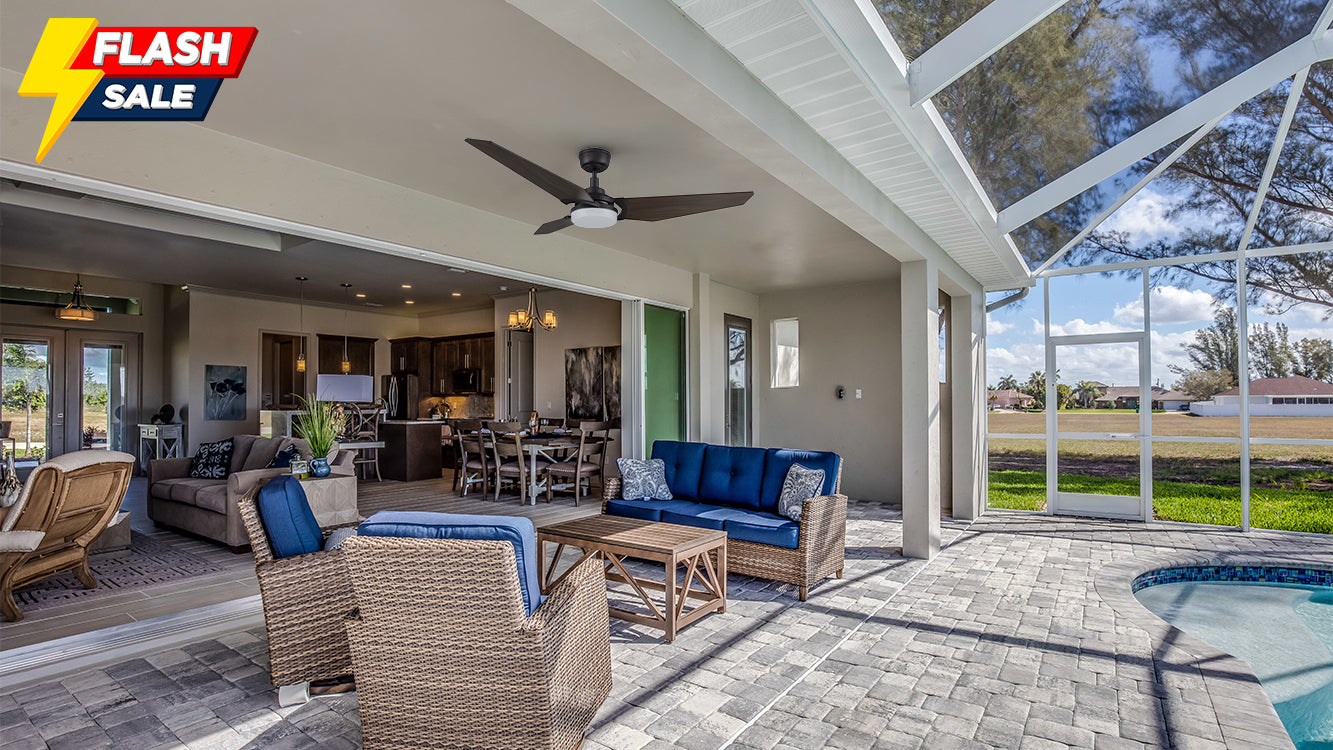 Indoor-outdoor living area with a black flush-mount smart ceiling fan with light and wooden blades hanging from the top. The patio features wicker chairs and a sofa with blue cushions, paired with a wooden coffee table. The background shows an indoor kitchen and dining area with light gray walls. In the distance, a green lawn and swimming pool are visible, with a transparent roof overhead and trees and blue sky in the background.