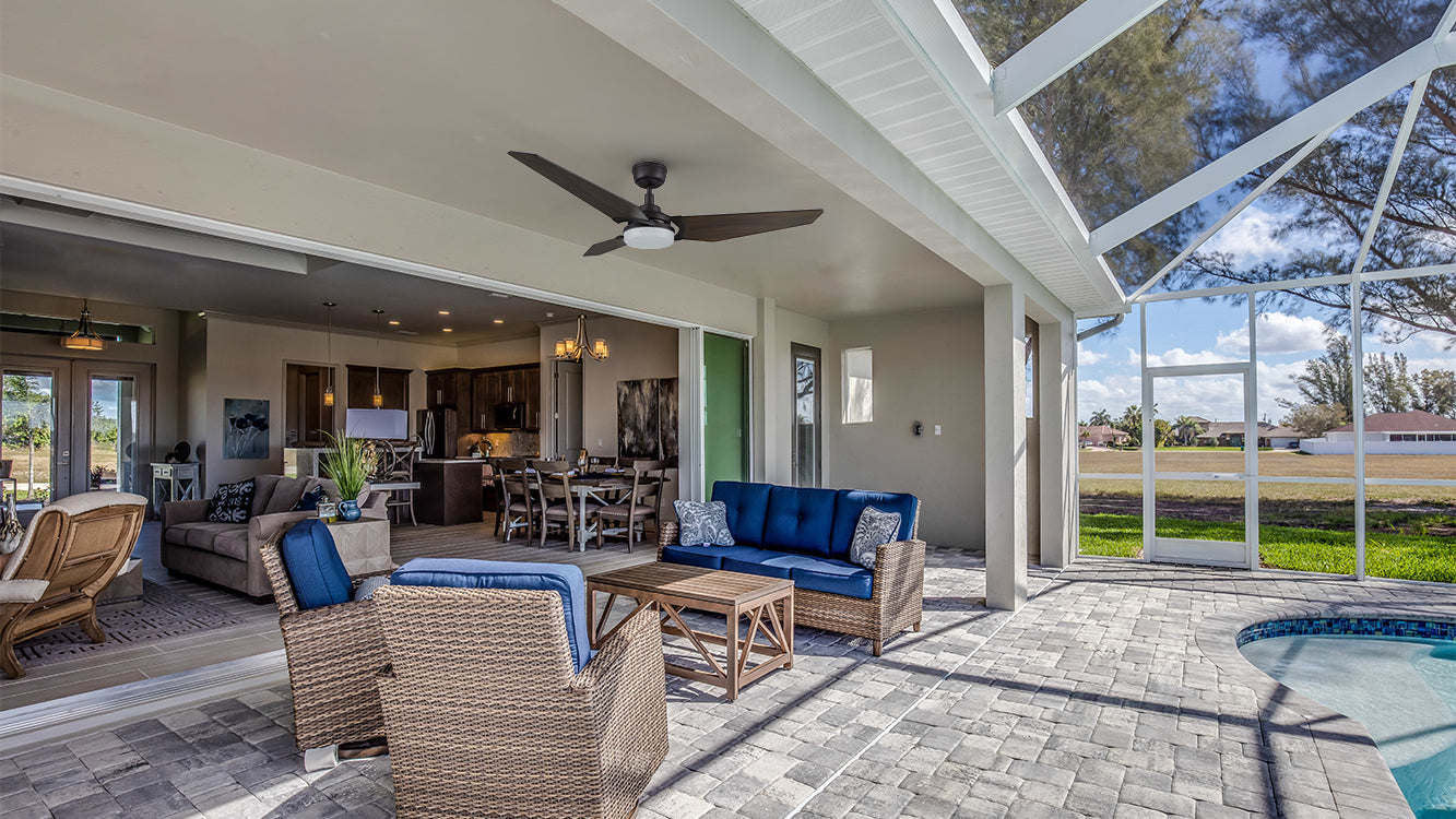 Indoor-outdoor living area with a black flush-mount smart ceiling fan with light and wooden blades hanging from the top. The patio features wicker chairs and a sofa with blue cushions, paired with a wooden coffee table. The background shows an indoor kitchen and dining area with light gray walls. In the distance, a green lawn and swimming pool are visible, with a transparent roof overhead and trees and blue sky in the background.