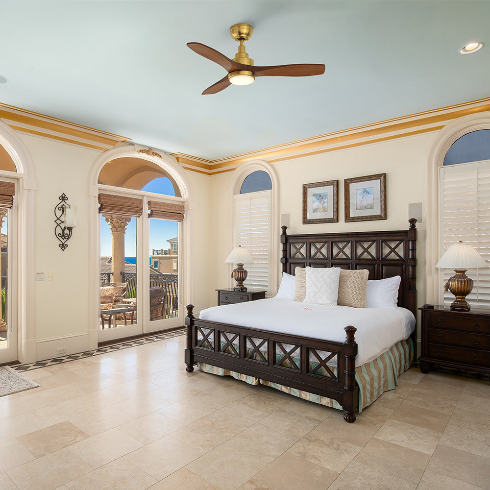 A luxurious bedroom with a large dark wood four-poster bed and marble floors, featuring arched glass doors leading to a balcony with an ocean view, and a Smafan Ultiem 48-inch wood ceiling fan in a gold finish with LED light mounted on the light blue ceiling. 