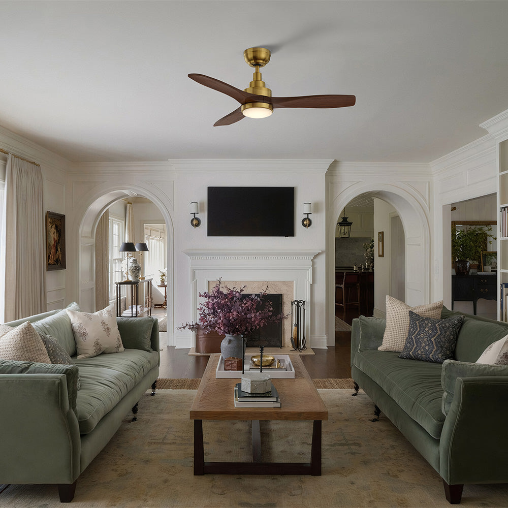 A living room with light walls and several arched doorways leading to other areas. The room contains two green velvet sofas facing each other across a rectangular wooden coffee table. A fireplace and a wall-mounted television are positioned centrally, above which hangs a gold and wood-finish Smafan Ultiem 48-inch ceiling fan with an LED light and remote. 