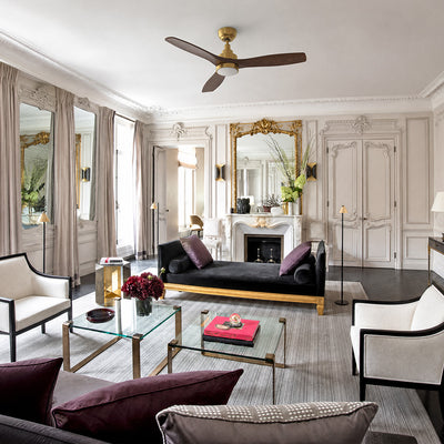 Smafan Ultiem 48 inch ceiling fan with LED light in a French country living room. The gold ceiling fan with three dark wood blades complements the elegant space featuring ornate wall moldings, a marble fireplace with a gilded mirror, black velvet seating, glass coffee tables, and classic French-inspired décor. #color_gold