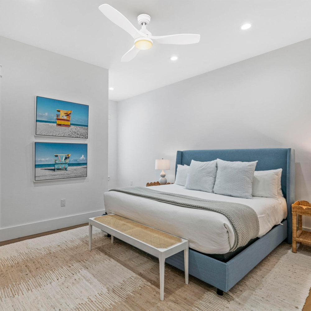 Minimalist bedroom with neutral bedding, nightstands, and coastal artwork; Ultiem 48-inch white ceiling fan with integrated LED positioned over the bed for quiet cooling and soft light. 