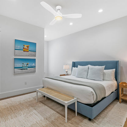 Minimalist bedroom with neutral bedding, nightstands, and coastal artwork; Ultiem 48-inch white ceiling fan with integrated LED positioned over the bed for quiet cooling and soft light. 