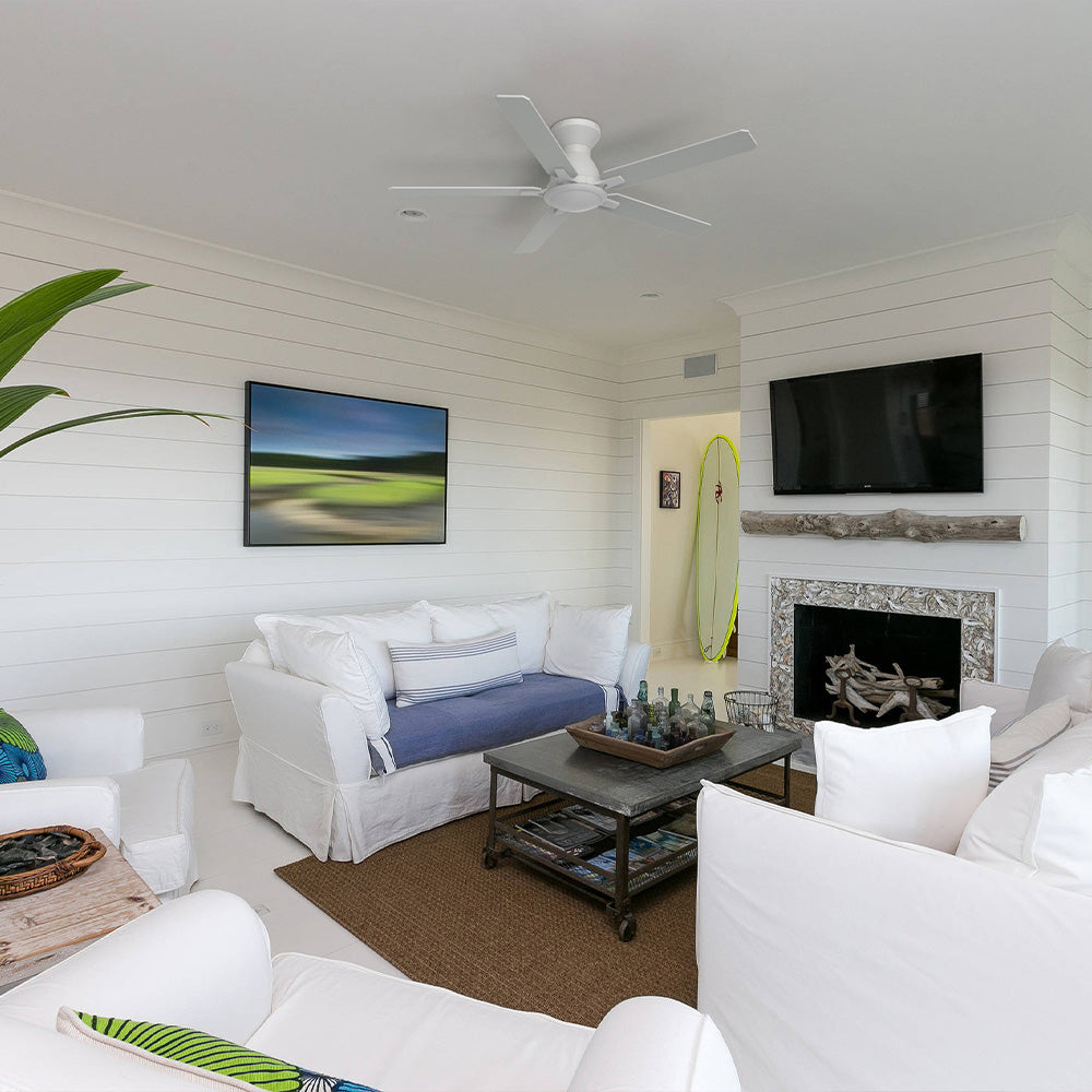 A bright, beach-themed living room featuring white shiplap walls, white slipcovered sofas and armchairs, a central fireplace with a natural wood mantelpiece, a wall-mounted TV, a dark industrial-style coffee table, and a green and yellow surfboard leaning in a nearby alcove. 