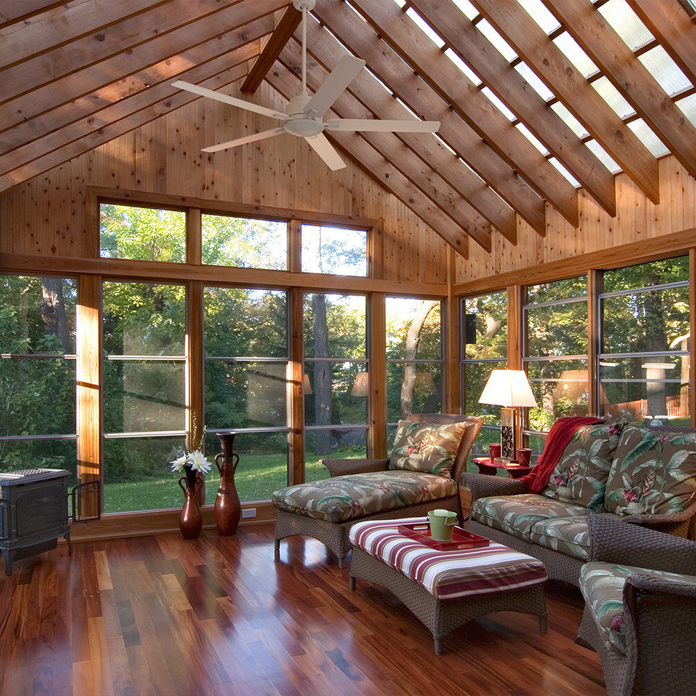 A spacious, sunlit sunroom with a high, exposed wood-beam vaulted ceiling and large windows overlooking a green yard. A white five-blade ceiling fan is mounted on a long downrod. The room features wicker furniture with white and patterned cushions, a striped ottoman, and dark wood flooring, evoking a farmhouse living room or rustic style. Smafan Warwick 60-inch ceiling fan for vaulted ceilings. 